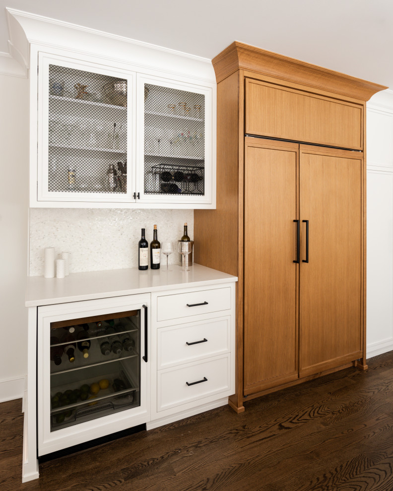 This is an example of a mid-sized transitional l-shaped home bar in New York with a drop-in sink, beaded inset cabinets, white cabinets, solid surface benchtops, white splashback, porcelain splashback, medium hardwood floors and white benchtop.