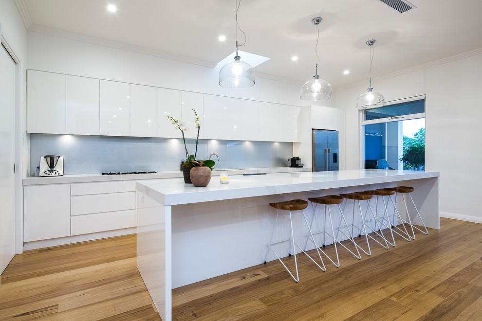 Inspiration for a contemporary galley kitchen in Adelaide with flat-panel cabinets, white cabinets, white splashback, glass sheet splashback, stainless steel appliances, medium hardwood floors, with island and brown floor.