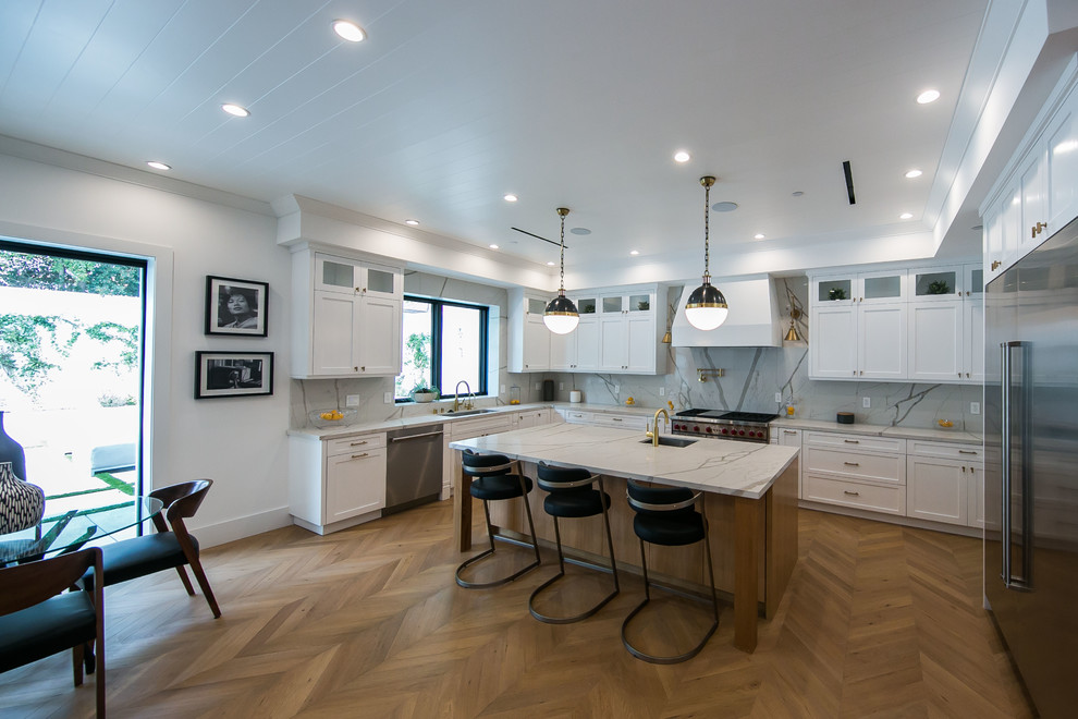 This is an example of a mid-sized country l-shaped eat-in kitchen in Los Angeles with a double-bowl sink, glass-front cabinets, white cabinets, quartz benchtops, beige splashback, stainless steel appliances, light hardwood floors, with island, brown floor and beige benchtop.