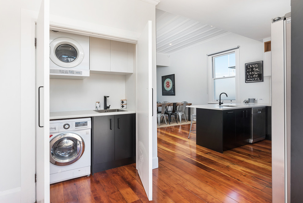 Inspiration for a mid-sized modern u-shaped laundry room in Auckland with an undermount sink, flat-panel cabinets, black cabinets, laminate benchtops, white splashback, ceramic splashback, medium hardwood floors and brown floor.