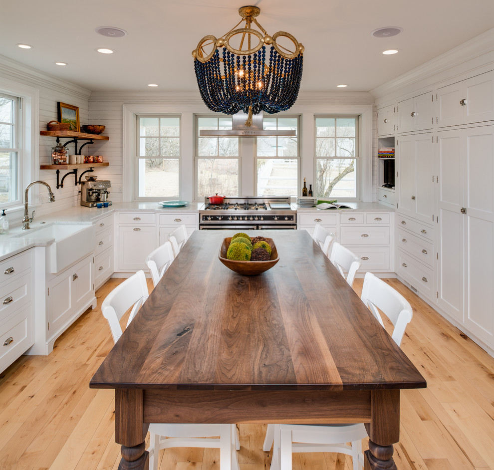 Mid-sized beach style u-shaped eat-in kitchen in Other with a farmhouse sink, white cabinets, marble benchtops, white splashback, black appliances, light hardwood floors, with island, beaded inset cabinets, window splashback and beige floor.