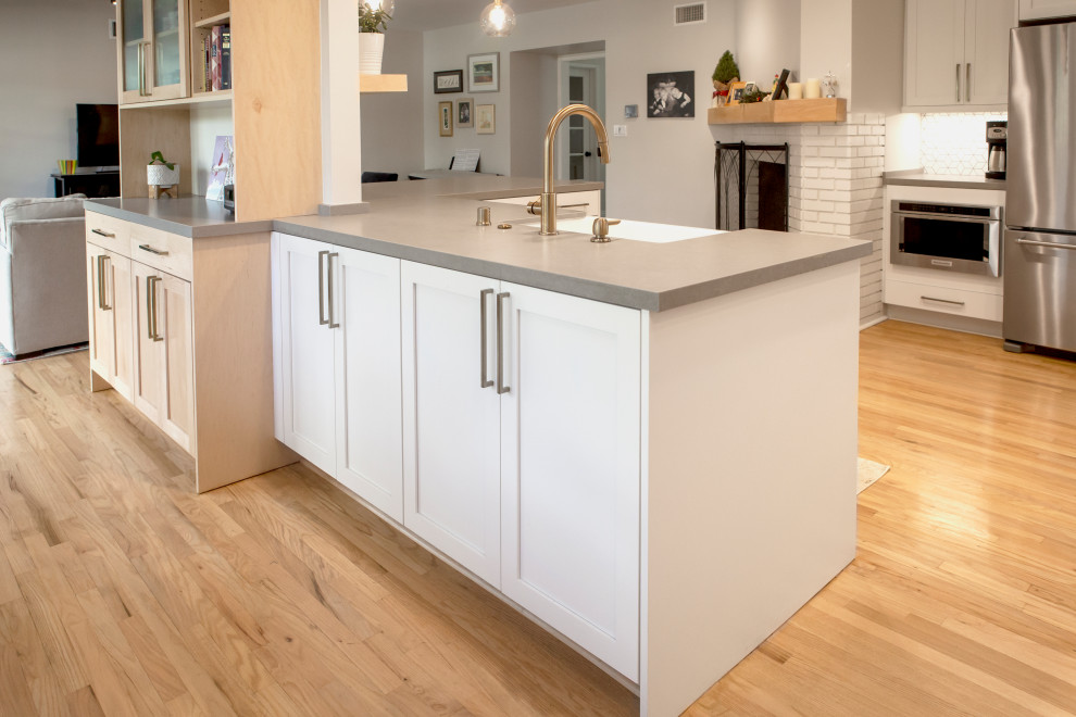 This is an example of a large midcentury l-shaped eat-in kitchen in Los Angeles with a farmhouse sink, shaker cabinets, white cabinets, quartz benchtops, white splashback, ceramic splashback, stainless steel appliances, light hardwood floors, a peninsula, grey benchtop and recessed.