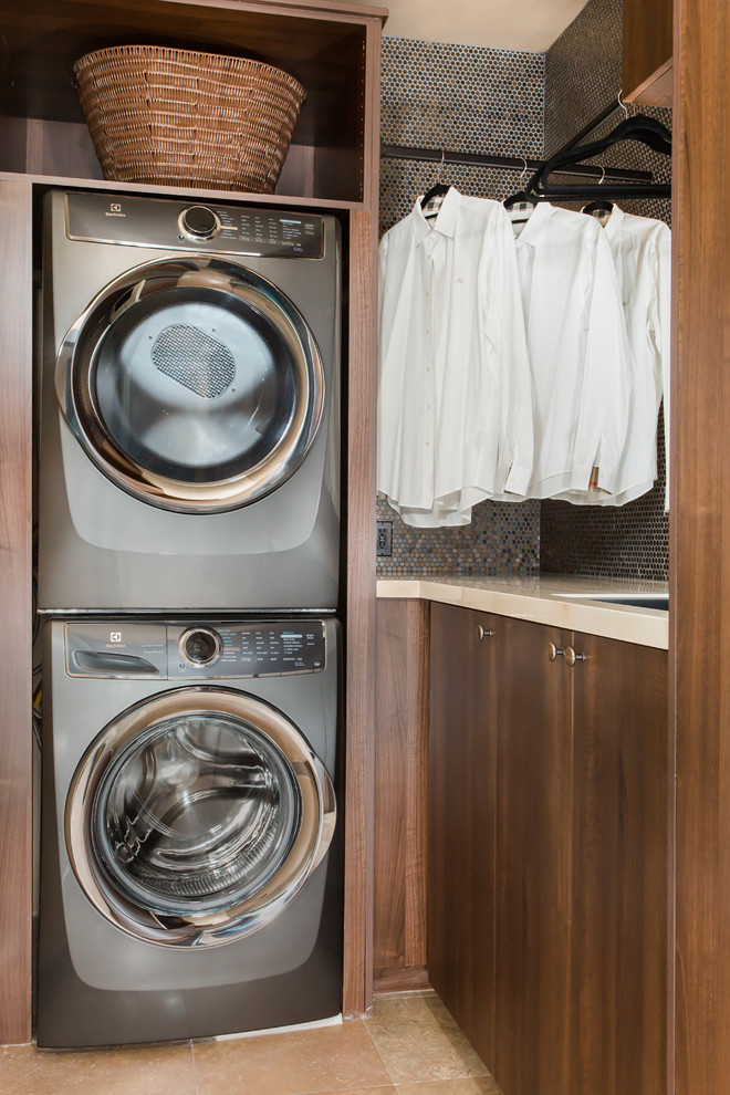 Inspiration for a mid-sized contemporary l-shaped dedicated laundry room in Los Angeles with an undermount sink, flat-panel cabinets, dark wood cabinets, solid surface benchtops, beige walls, travertine floors, a stacked washer and dryer and beige floor.