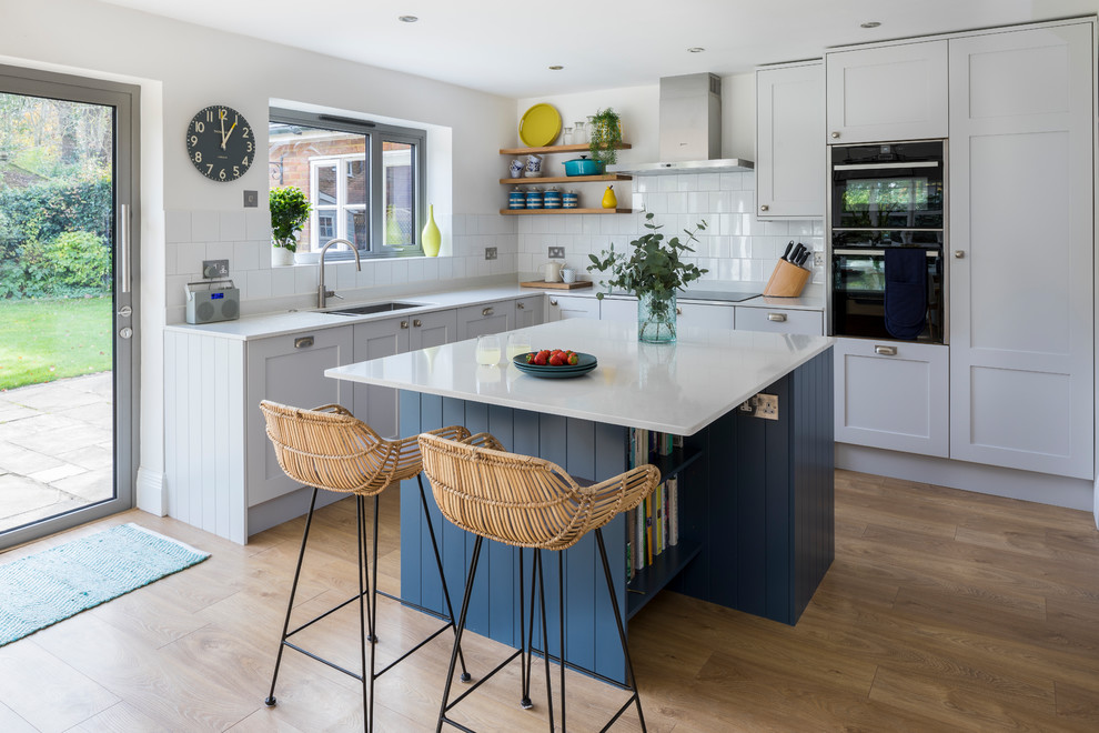 Inspiration for a mid-sized transitional l-shaped kitchen in Surrey with an undermount sink, shaker cabinets, solid surface benchtops, white splashback, porcelain splashback, with island, white benchtop, grey cabinets, black appliances, medium hardwood floors and brown floor.