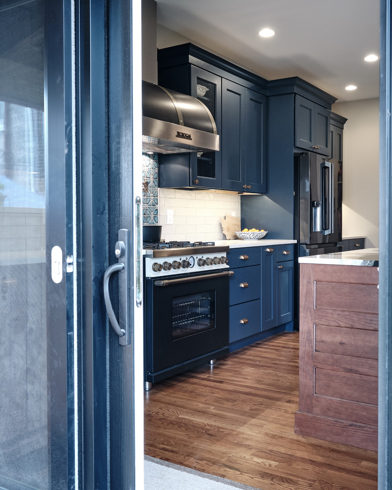 Design ideas for a large eclectic galley open plan kitchen in Philadelphia with a farmhouse sink, recessed-panel cabinets, blue cabinets, quartzite benchtops, grey splashback, ceramic splashback, black appliances, medium hardwood floors, with island, brown floor and multi-coloured benchtop.