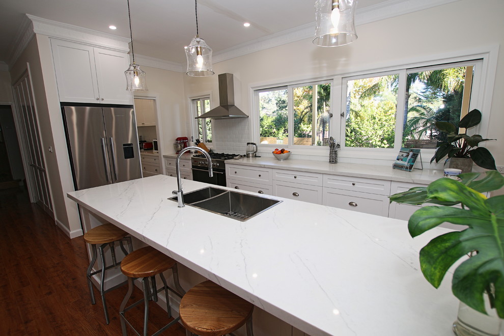 Inspiration for a large transitional galley open plan kitchen in Sydney with a double-bowl sink, shaker cabinets, white cabinets, quartz benchtops, white splashback, subway tile splashback, stainless steel appliances, medium hardwood floors, with island, brown floor and white benchtop.