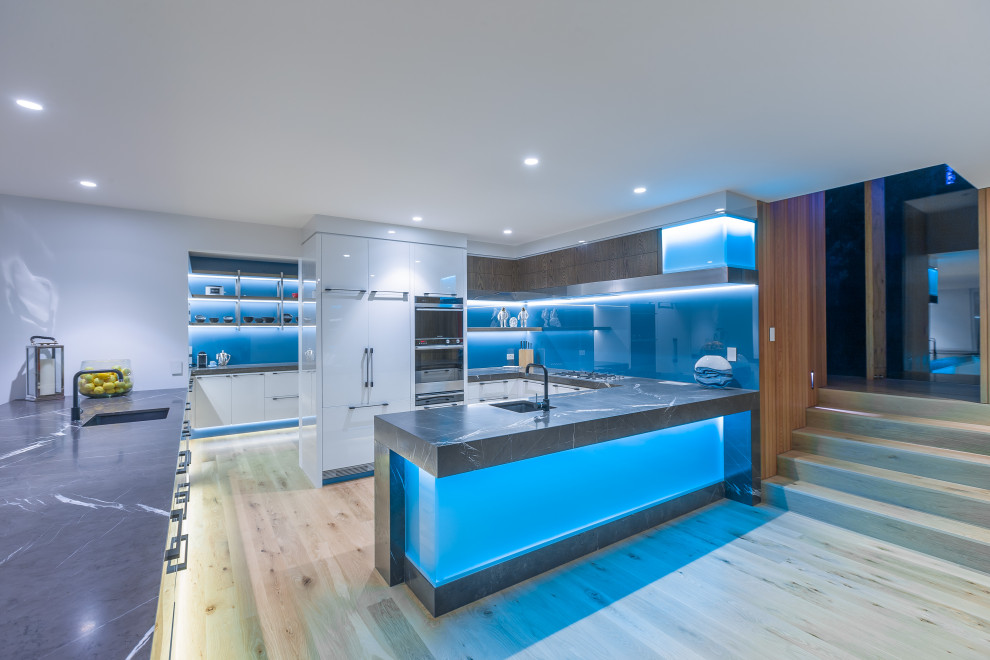 Design ideas for an expansive contemporary l-shaped open plan kitchen in Auckland with an undermount sink, flat-panel cabinets, white cabinets, granite benchtops, blue splashback, glass sheet splashback, stainless steel appliances, light hardwood floors, grey benchtop, a peninsula and beige floor.