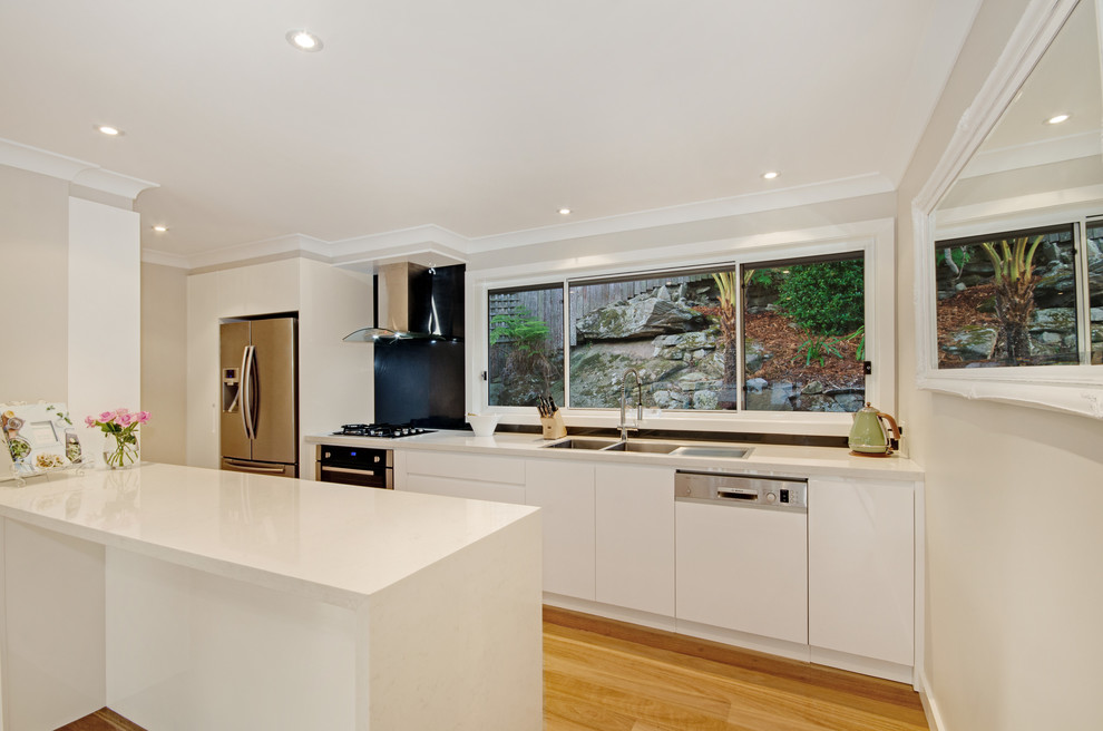 Design ideas for a mid-sized modern l-shaped kitchen pantry in Sydney with a double-bowl sink, flat-panel cabinets, white cabinets, marble benchtops, blue splashback, glass sheet splashback, stainless steel appliances and light hardwood floors.