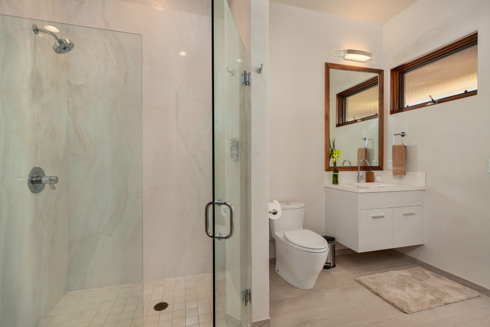 This is an example of a mid-sized tropical master wet room bathroom in Hawaii with porcelain floors, flat-panel cabinets, white cabinets, a one-piece toilet, white tile, stone slab, white walls, a drop-in sink, engineered quartz benchtops and a hinged shower door.