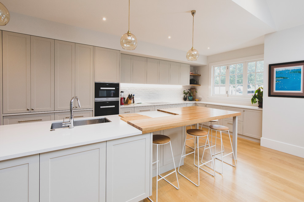 Large country l-shaped kitchen in Auckland with shaker cabinets, white splashback, ceramic splashback, stainless steel appliances, light hardwood floors, with island, white benchtop, an undermount sink, grey cabinets and beige floor.