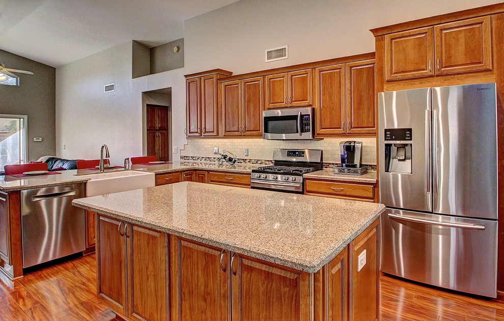 Inspiration for a mid-sized l-shaped medium tone wood floor eat-in kitchen remodel in Phoenix with a farmhouse sink, raised-panel cabinets, medium tone wood cabinets, beige backsplash, stone tile backsplash, stainless steel appliances and an island