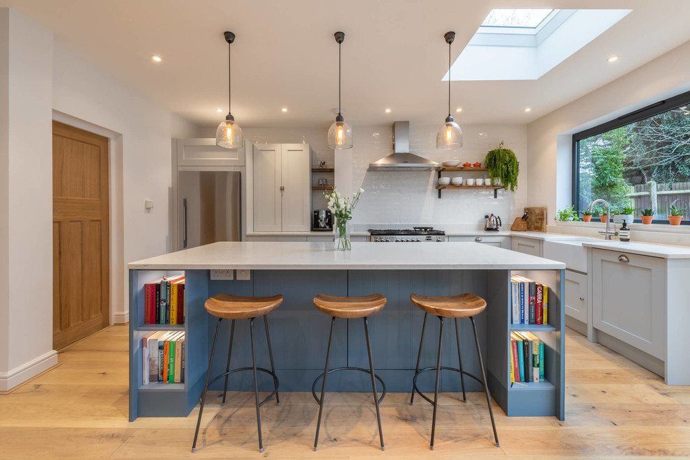 This is an example of a transitional l-shaped kitchen in Sussex with a farmhouse sink, shaker cabinets, grey cabinets, white splashback, subway tile splashback, light hardwood floors, with island, beige floor and white benchtop.
