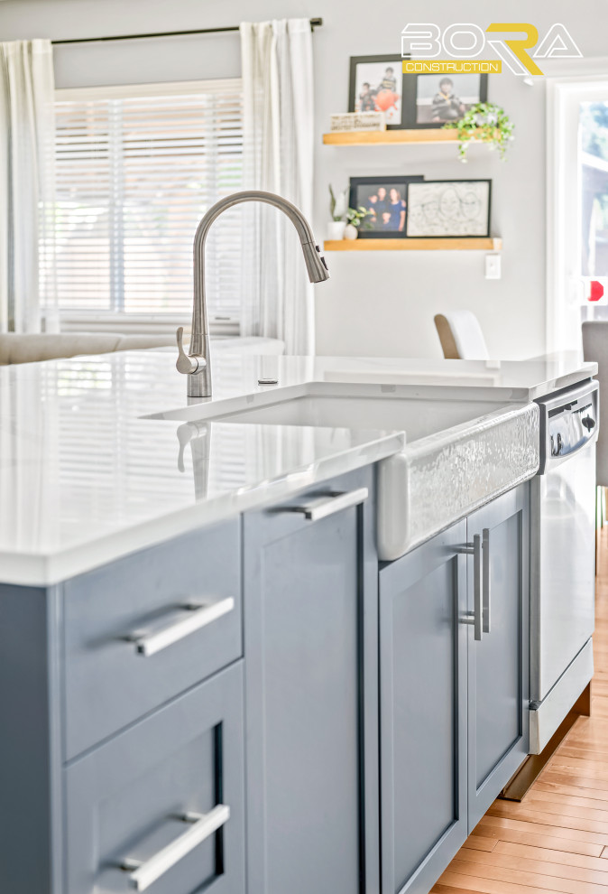 Example of a mid-sized single-wall light wood floor and brown floor eat-in kitchen design in Seattle with a farmhouse sink, shaker cabinets, white cabinets, quartz countertops, white backsplash, quartz backsplash, stainless steel appliances, an island and white countertops