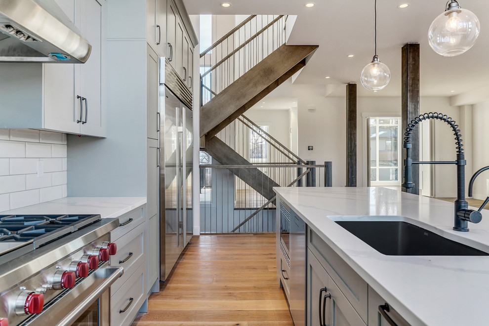 Inspiration for a mid-sized transitional l-shaped medium tone wood floor and brown floor eat-in kitchen remodel in Calgary with a single-bowl sink, shaker cabinets, gray cabinets, quartzite countertops, white backsplash, ceramic backsplash, stainless steel appliances, an island and white countertops