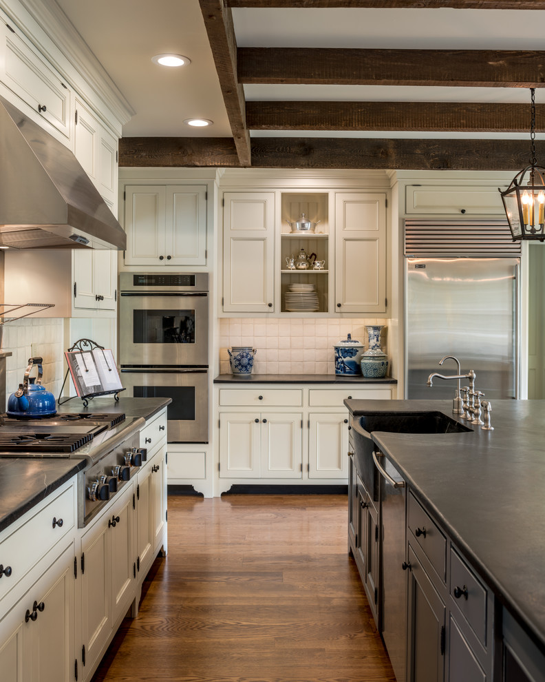This is an example of an expansive traditional u-shaped separate kitchen in Philadelphia with a farmhouse sink, beaded inset cabinets, beige cabinets, soapstone benchtops, beige splashback, stainless steel appliances, medium hardwood floors, with island, brown floor and black benchtop.