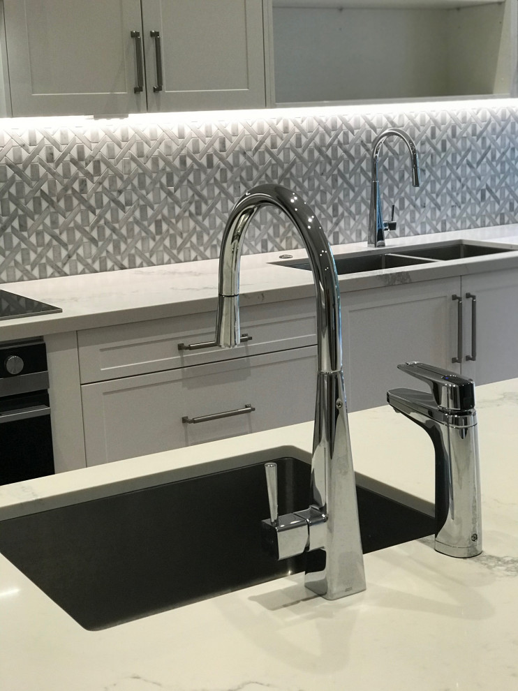 This is an example of a large modern l-shaped kitchen pantry in Sydney with an undermount sink, shaker cabinets, white cabinets, quartz benchtops, grey splashback, mosaic tile splashback, black appliances, medium hardwood floors, with island, brown floor and white benchtop.