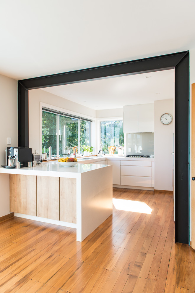 Inspiration for a mid-sized scandinavian u-shaped kitchen pantry in Christchurch with an undermount sink, flat-panel cabinets, white cabinets, solid surface benchtops, glass sheet splashback, white appliances, medium hardwood floors, a peninsula and brown floor.