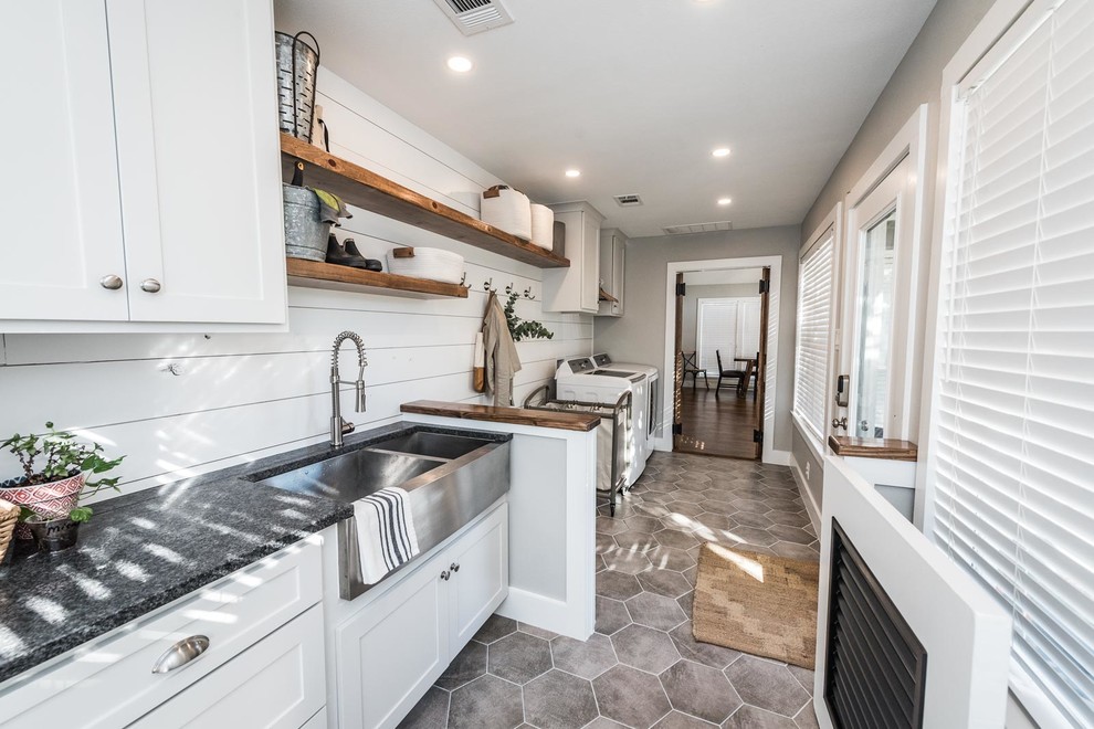 Photo of a large country galley dedicated laundry room in Dallas with a farmhouse sink, shaker cabinets, white cabinets, granite benchtops, white walls, ceramic floors, a side-by-side washer and dryer, grey floor and black benchtop.