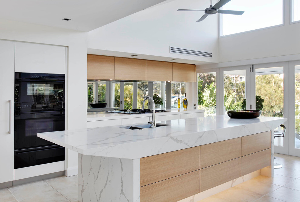 Photo of a large modern l-shaped open plan kitchen in Sydney with an undermount sink, flat-panel cabinets, white cabinets, quartz benchtops, grey splashback, mirror splashback, black appliances, limestone floors and with island.