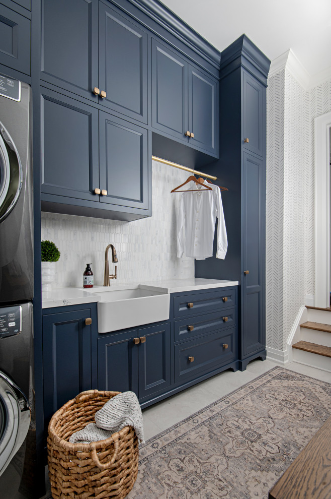 Traditional single-wall dedicated laundry room in Toronto with recessed-panel cabinets, blue cabinets, white benchtop, a farmhouse sink, white splashback, a stacked washer and dryer and wallpaper.