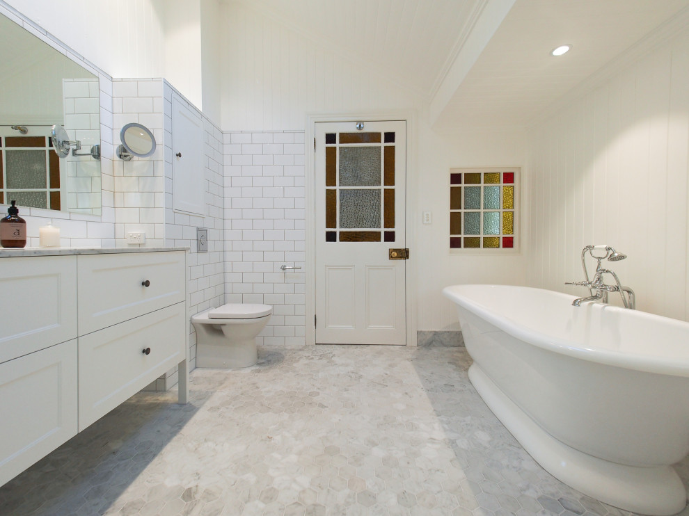 Large traditional master bathroom with beaded inset cabinets, white cabinets, a freestanding tub, a curbless shower, a one-piece toilet, white tile, porcelain tile, white walls, marble floors, an undermount sink and marble benchtops.