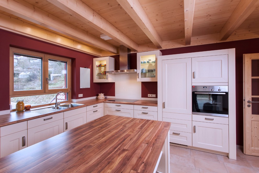 Photo of a large country l-shaped separate kitchen in Stuttgart with a drop-in sink, white cabinets, wood benchtops, red splashback, terra-cotta floors, with island, beaded inset cabinets, panelled appliances and brown benchtop.