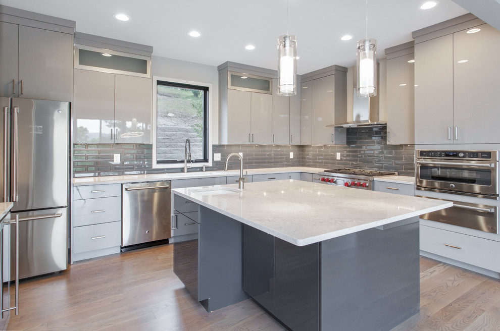 Large modern l-shaped kitchen pantry in Cincinnati with a drop-in sink, grey cabinets, quartzite benchtops, grey splashback, glass tile splashback, stainless steel appliances, light hardwood floors, with island, brown floor and white benchtop.