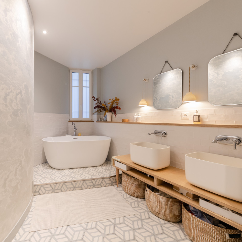 Photo of a large scandinavian master bathroom in Paris with open cabinets, light wood cabinets, a freestanding tub, a shower/bathtub combo, white tile, terra-cotta tile, grey walls, cement tiles, a vessel sink, wood benchtops, grey floor, an open shower, beige benchtops, a double vanity, a freestanding vanity and wallpaper.