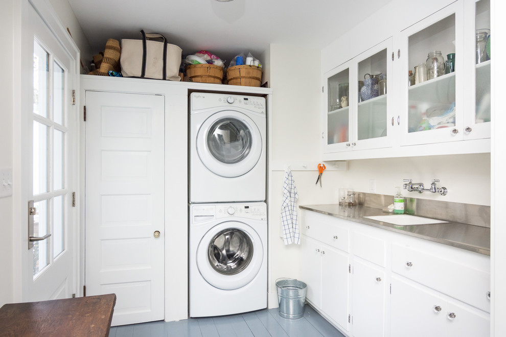 Inspiration for a large beach style l-shaped laundry room in Detroit with an undermount sink, glass-front cabinets, white cabinets, stainless steel benchtops, white walls, painted wood floors and a stacked washer and dryer.