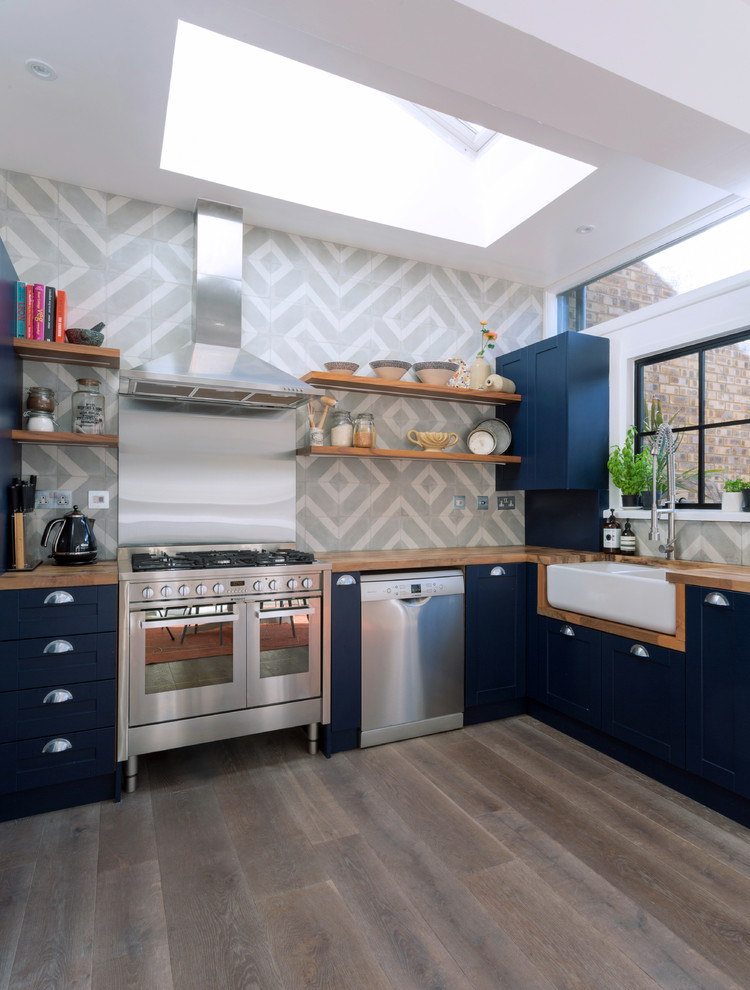 This is an example of a transitional open plan kitchen in London with a farmhouse sink, recessed-panel cabinets, blue cabinets, wood benchtops, grey splashback, cement tile splashback, stainless steel appliances and light hardwood floors.