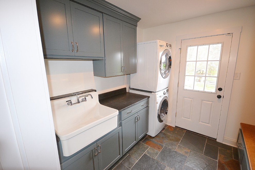 Design ideas for a mid-sized transitional single-wall utility room in Philadelphia with a farmhouse sink, flat-panel cabinets, wood benchtops, white walls, ceramic floors, a stacked washer and dryer and grey cabinets.