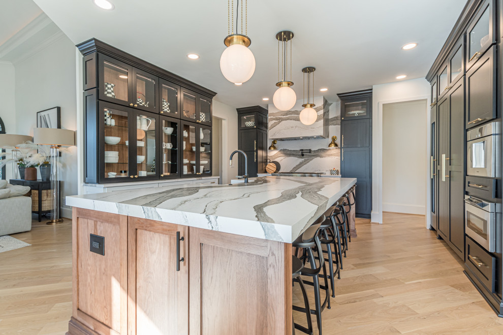 This is an example of a large transitional u-shaped eat-in kitchen in Atlanta with a farmhouse sink, recessed-panel cabinets, black cabinets, quartz benchtops, white splashback, engineered quartz splashback, stainless steel appliances, light hardwood floors, with island, brown floor and white benchtop.