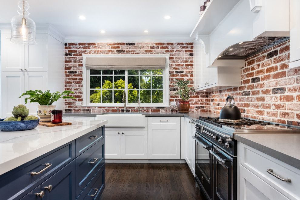Inspiration for a mid-sized country l-shaped open plan kitchen in Los Angeles with a farmhouse sink, shaker cabinets, white cabinets, quartz benchtops, multi-coloured splashback, brick splashback, black appliances, dark hardwood floors, with island, brown floor and grey benchtop.