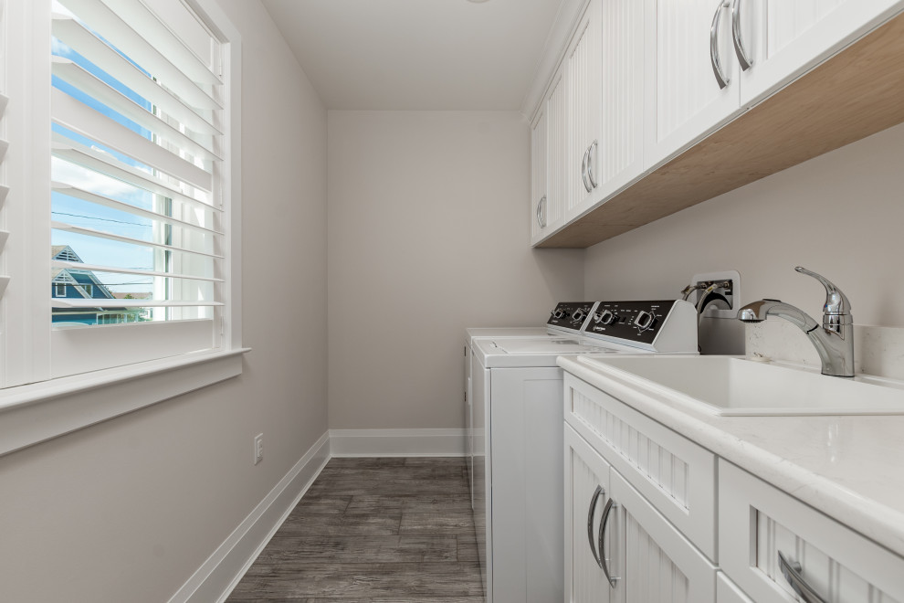 Photo of a small beach style galley dedicated laundry room in Other with a drop-in sink, beaded inset cabinets, white cabinets, solid surface benchtops, grey walls, porcelain floors, a side-by-side washer and dryer, grey floor and white benchtop.