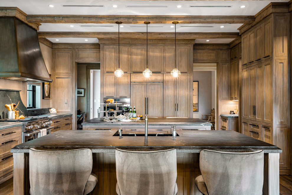 This is an example of an expansive world-inspired grey and brown kitchen/diner in Miami with recessed-panel cabinets, medium wood cabinets, soapstone worktops, stainless steel appliances, dark hardwood flooring, multiple islands, brown floors, exposed beams, feature lighting, a belfast sink and green worktops.
