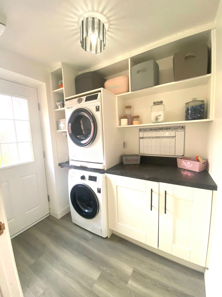 Imagen de lavadero multiusos y lineal de tamaño medio con armarios estilo shaker, puertas de armario blancas, lavadora y secadora apiladas y encimeras negras