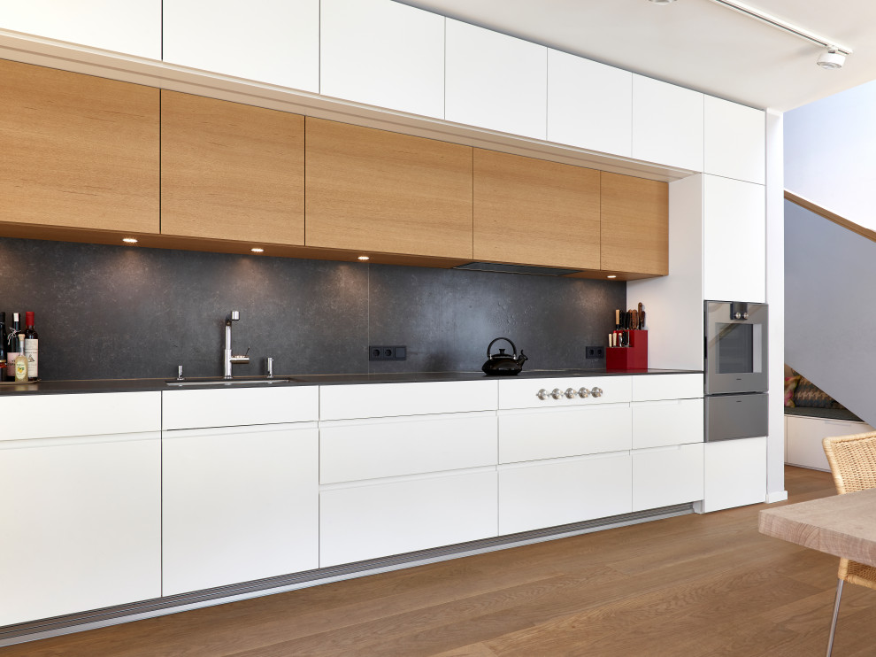 Design ideas for a large contemporary single-wall open plan kitchen in Munich with an integrated sink, flat-panel cabinets, white cabinets, concrete benchtops, black splashback, metal splashback, stainless steel appliances, medium hardwood floors, no island, brown floor and black benchtop.
