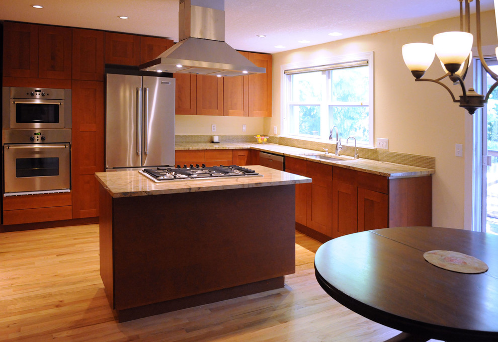 Mid-sized transitional l-shaped eat-in kitchen in Portland with a double-bowl sink, medium wood cabinets, granite benchtops, glass tile splashback, stainless steel appliances, light hardwood floors, with island, brown floor, multi-coloured splashback, multi-coloured benchtop and shaker cabinets.