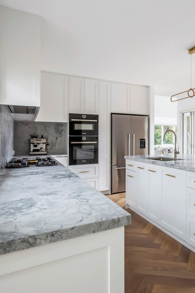 This is an example of a large beach style l-shaped open plan kitchen in Sydney with an undermount sink, shaker cabinets, white cabinets, marble benchtops, grey splashback, marble splashback, stainless steel appliances, light hardwood floors, with island, multi-coloured floor, grey benchtop and coffered.
