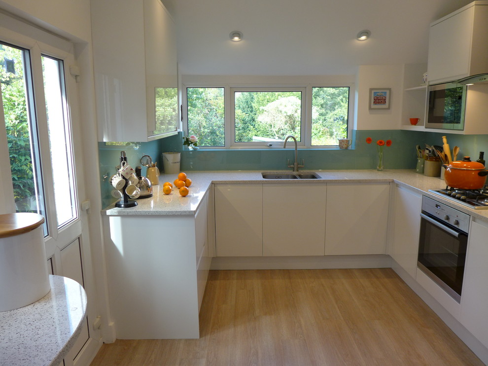 This is an example of a small modern u-shaped enclosed kitchen with a built-in sink, flat-panel cabinets, white cabinets, quartz worktops, blue splashback, glass sheet splashback, stainless steel appliances and vinyl flooring.