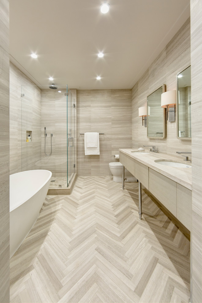 This is an example of a large contemporary master bathroom in New York with an undermount sink, flat-panel cabinets, light wood cabinets, limestone benchtops, a freestanding tub, a corner shower, a one-piece toilet, stone tile, brown walls, limestone floors and beige tile.