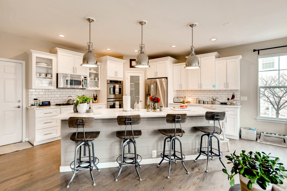 Inspiration for an expansive country galley eat-in kitchen in Denver with a farmhouse sink, shaker cabinets, white cabinets, quartz benchtops, white splashback, ceramic splashback, stainless steel appliances, light hardwood floors, with island, brown floor and white benchtop.