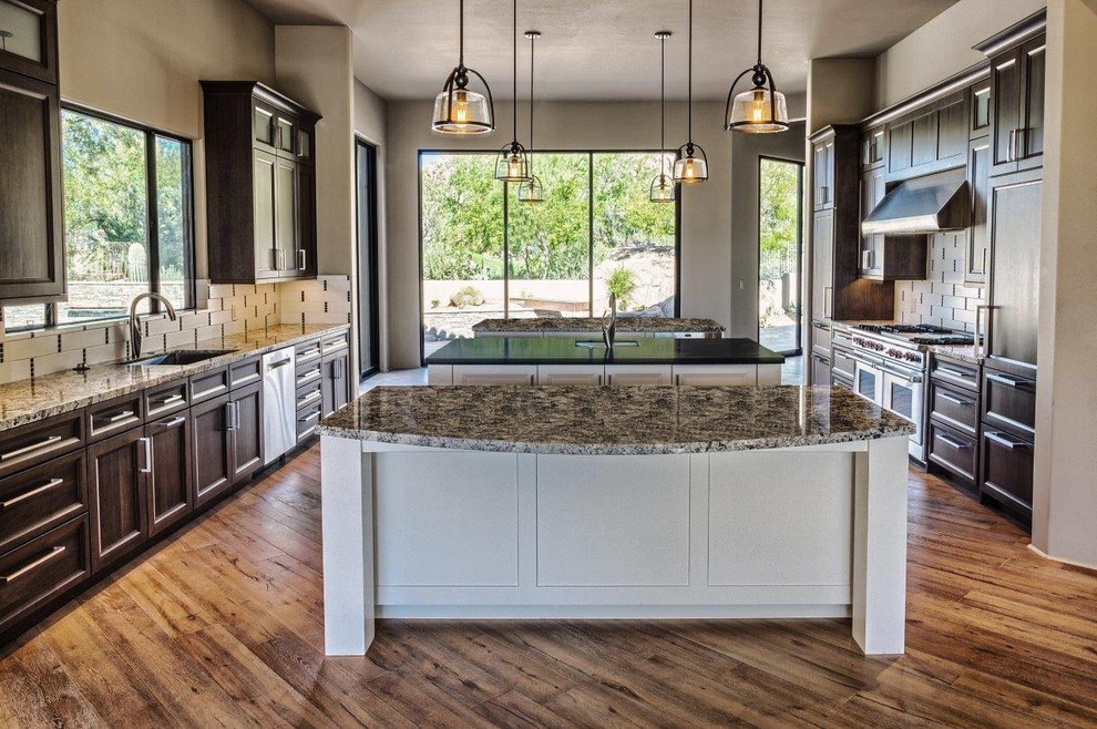 Kitchen - large transitional medium tone wood floor kitchen idea in Phoenix with dark wood cabinets, granite countertops, paneled appliances, two islands, an undermount sink, recessed-panel cabinets, white backsplash and porcelain backsplash