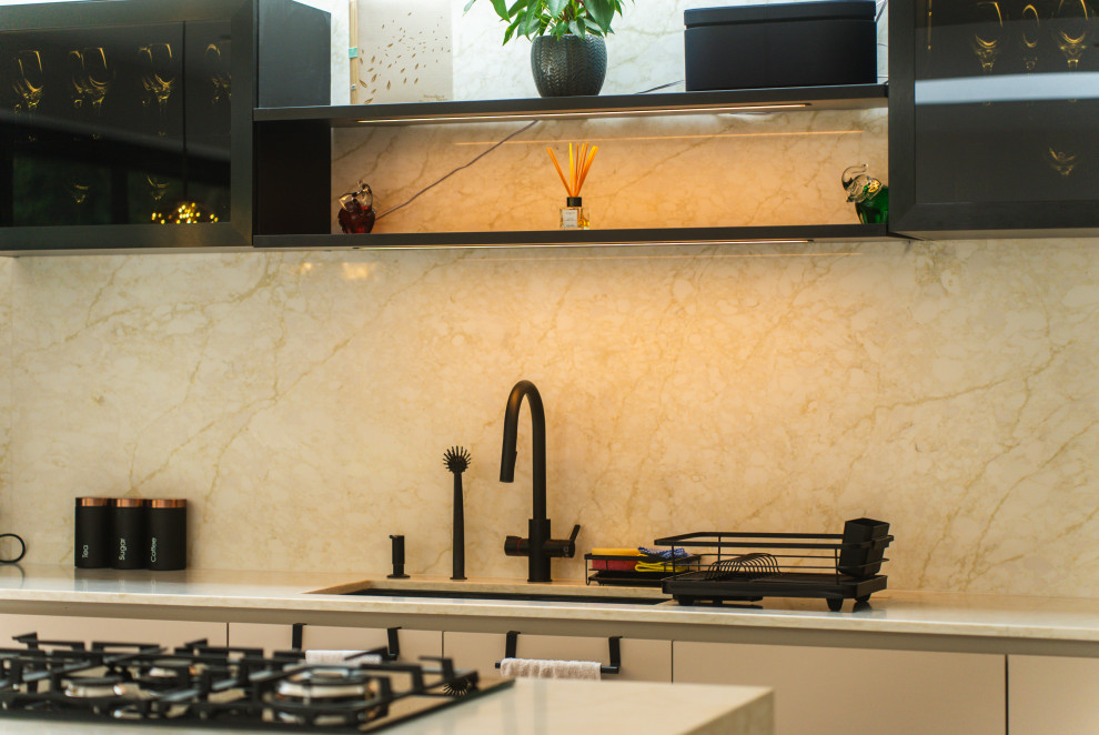 Photo of a medium sized cream and black single-wall open plan kitchen in London with beige cabinets, quartz worktops, beige splashback, engineered quartz splashback, black appliances, porcelain flooring, an island, grey floors, beige worktops and feature lighting.