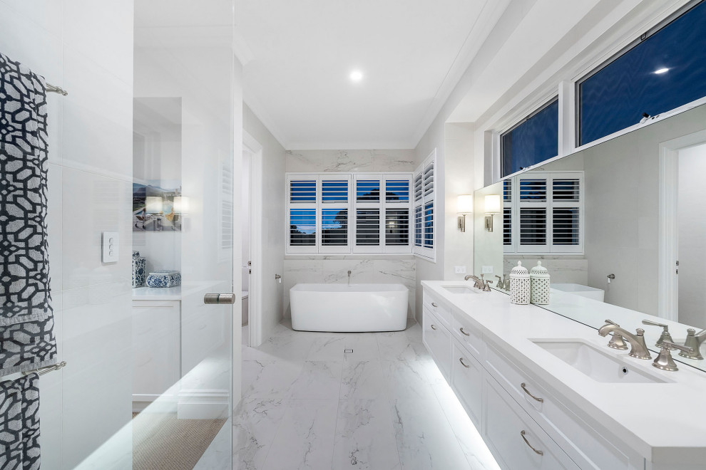 Large beach style master bathroom in Perth with recessed-panel cabinets, white cabinets, a freestanding tub, a curbless shower, a wall-mount toilet, white tile, porcelain tile, white walls, porcelain floors, an undermount sink, solid surface benchtops, white floor, a hinged shower door and white benchtops.