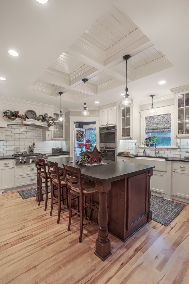 Inspiration for a traditional l-shaped kitchen in Milwaukee with a farmhouse sink, shaker cabinets, white cabinets, white splashback, subway tile splashback, stainless steel appliances, light hardwood floors and with island.
