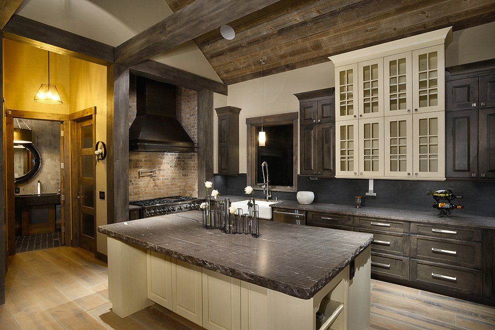 Inspiration for a large country l-shaped open plan kitchen in Denver with a farmhouse sink, recessed-panel cabinets, distressed cabinets, soapstone benchtops, black splashback, stone slab splashback, stainless steel appliances, medium hardwood floors, with island and brown floor.