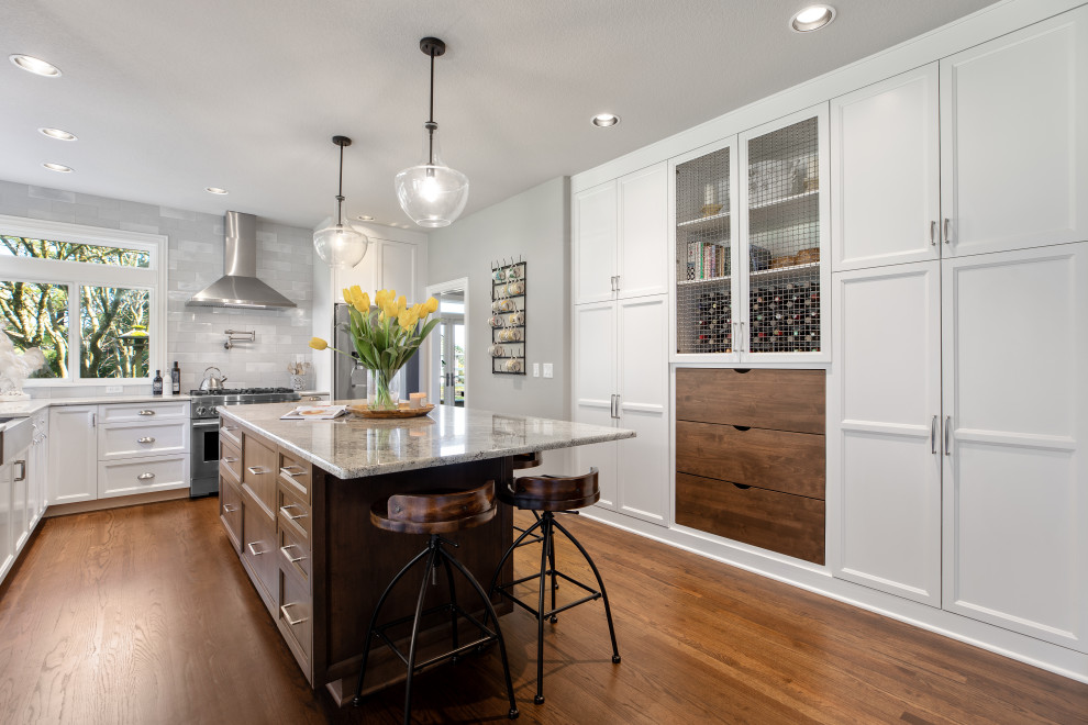 Large transitional u-shaped eat-in kitchen in Portland with a farmhouse sink, recessed-panel cabinets, white cabinets, granite benchtops, grey splashback, porcelain splashback, stainless steel appliances, dark hardwood floors, with island, brown floor and grey benchtop.