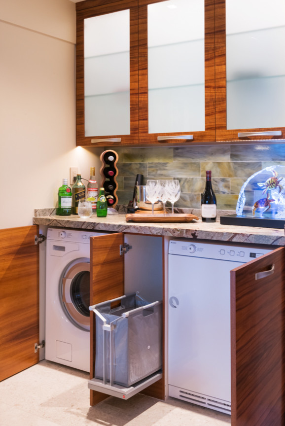 This is an example of a transitional l-shaped utility room in Hawaii with flat-panel cabinets, medium wood cabinets, marble benchtops, beige walls, limestone floors and a concealed washer and dryer.