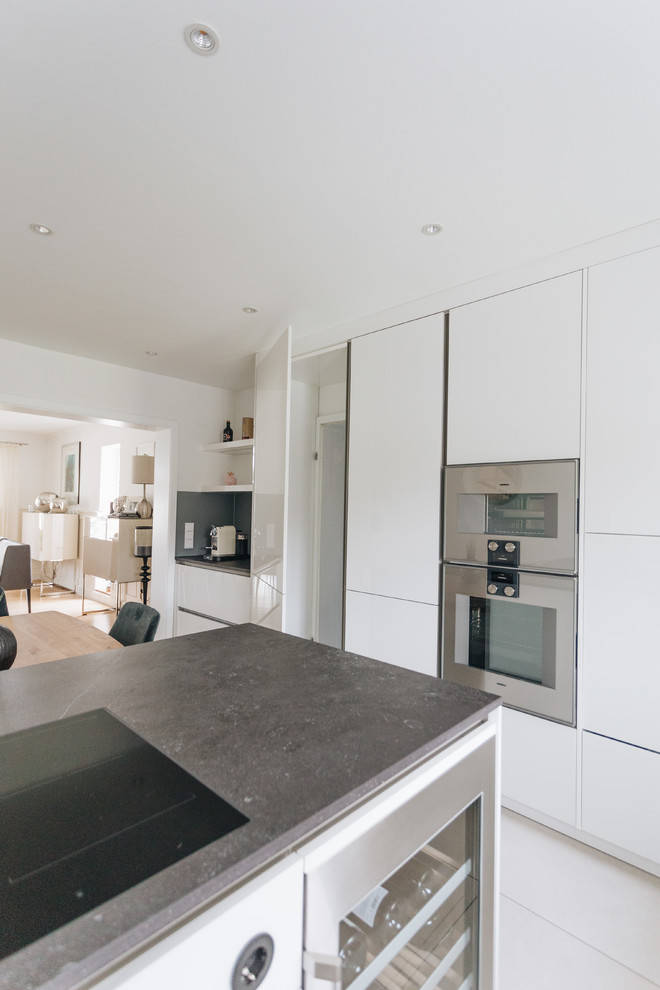 Large contemporary l-shaped open plan kitchen in Other with an undermount sink, flat-panel cabinets, white cabinets, granite benchtops, grey splashback, glass sheet splashback, stainless steel appliances, ceramic floors, with island, white floor and grey benchtop.